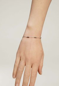 Pulsera de cadena de plata con pequeños eslabones ovalados y tres segmentos rectangulares con acentos oscuros, llevada en una mano.