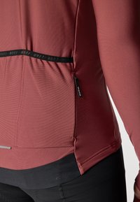 Bordeaux sporttop met een textuurontwerp, lange mouwen, een zwarte ritszak en een reflecterende accent nabij de zoom. Gecombineerd met zwarte onderkleding.