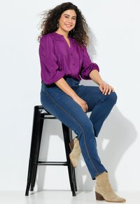 Fioletowa bluzka z bufkami, zapięta na guziki z przodu; zestawiona z ciemnoniebieskimi dżinsami z wysokim stanem i jasnobeżowymi zamszowymi botkami za kostkę. Czarny taboret.