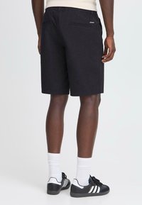 Shorts noirs en coton avec une taille élastique, des poches latérales et une poche arrière. Portés avec des baskets noires et blanches et des chaussettes blanches.