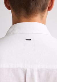 Witte linnen shirt met een klassieke kraag, een rugplooitje en een klein zwart label boven de kraag. De stof heeft een textuurafwerking.