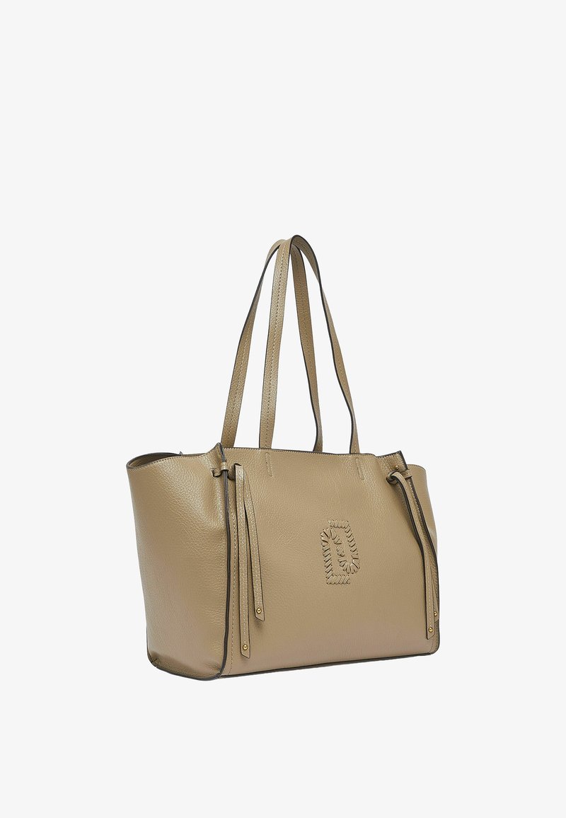 Sac fourre-tout en cuir beige avec deux bandoulières, pampilles décoratives sur les côtés et un emblème cousu à l'avant, posé sur un fond blanc.