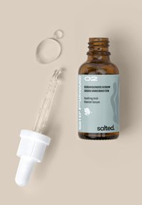 Bouteille en verre brun avec une étiquette verte, un capuchon compte-gouttes blanc, contenant un sérum clair ; une goutte de sérum à côté de la bouteille sur une surface beige.