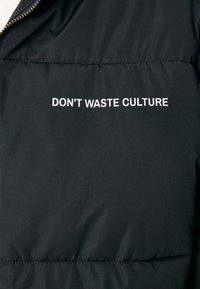 Crna puf jakna s mat površinom, sa bijelim izvezenim tekstom "NE TROŠITE KULTURU" preko prsnog dijela.