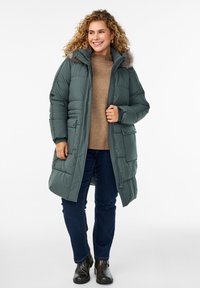 Frau mit lockigem Haar, die einen grünen gepolsterten Mantel mit Fellkapuze, einen beigen Pullover, dunkle Jeans und schwarze Stiefeletten trägt, lächelt und steht.