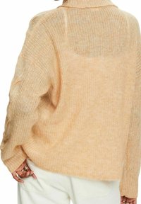 Personne portant un pull ample beige en maille côtelée avec des boutons en écaille sur la manche, associé à un pantalon blanc.