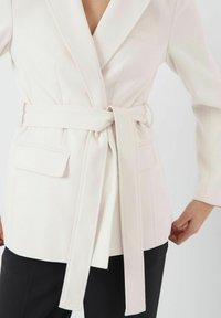 Lyserød skræddersyet blazer med bælte i taljen, snitlapeller og to forlommer. Glat stof med en struktureret design.