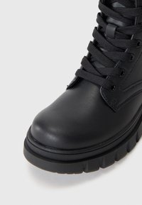 Botte de cheville en cuir noir avec un bout arrondi, une surface texturée, des lacets plats et une semelle en caoutchouc épaisse et striée. Design minimaliste.