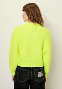 Neon żółty sweter dzianinowy o prążkowanej strukturze, zaokrąglonym dekolcie i opadających ramionach, zestawiony z czarnymi dżinsami z tylnymi kieszeniami.