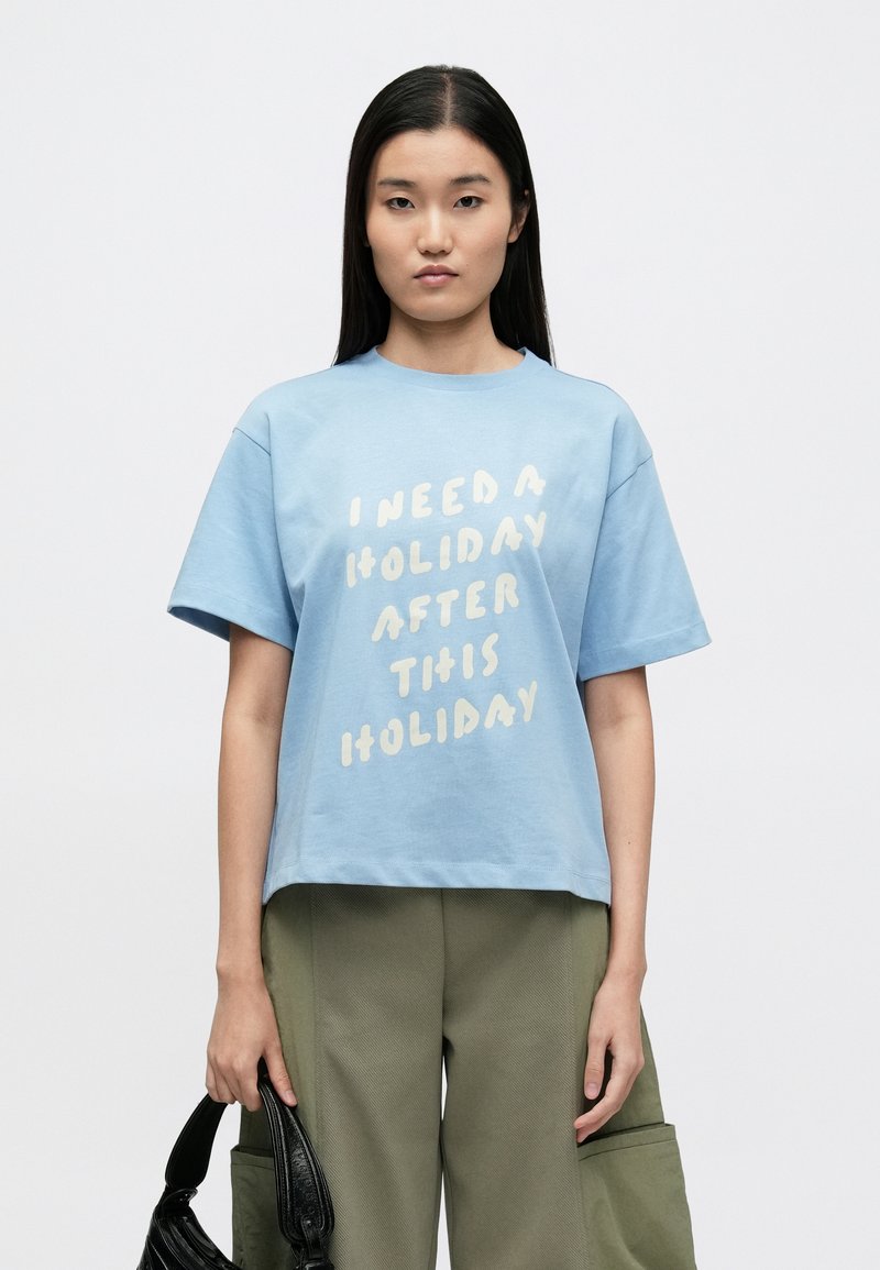 Ανοικτό μπλε βαμβακερό μπλουζάκι με κοντά μανίκια και στρογγυλή λαιμόκοψη. Διαθέτει μεγάλο μπεζ κείμενο που γράφει, «ΧΡΕΙΑΖΟΜΑΙ ΔΙΑΚΟΠΕΣ ΜΕΤΑ ΑΠΟ ΑΥΤΕΣ ΤΙΣ ΔΙΑΚΟΠΕΣ.»