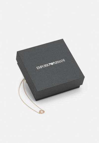 Caixa de joias preta com o logótipo prateado "EMPORIO ARMANI". Uma corrente dourada com um pendente circular sai da borda da caixa.