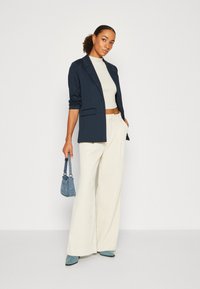 Blazer marine sur un top court crème, associé à un pantalon large ivoire. Les accessoires incluent un petit sac à main bleu et des chaussures teal.
