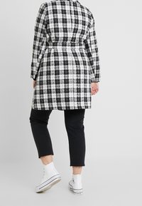 Abrigo largo a cuadros blanco y negro con cintura ajustada, combinado con jeans recortados negros y zapatillas de deporte blancas de caña alta. Textura suave.