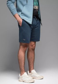 Niebieskie szorty dresowe wykonane z miękkiego materiału, z elastycznym paskiem w talii i bocznymi kieszeniami. Noszone z jasnoniebieską bluzą z kapturem i beżowymi trampkami.