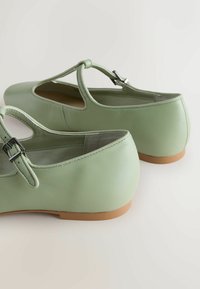 Paari helerohelisi lamedaid Mary Jane kingi, millel on pandlaga rihmad ja beežid kummist tallad, valgel taustal.