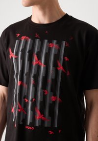 Schwarzes Baumwoll-T-Shirt mit einem Grafikdesign von roten Vögeln und vertikalen grauen Linien. Enthält ein Logodetail am unteren Rand.