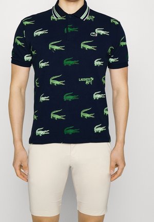 Granatowa koszulka polo z zielonymi wzorami krokodyli oraz tekstem "Lacoste Golf", noszona z beżowymi szortami na stojącej osobie.