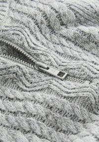 Tejido de suéter de punto gris claro con un patrón texturizado, que presenta una cremallera plateada a lo largo de la costura. El detalle en primer plano resalta el tejido.