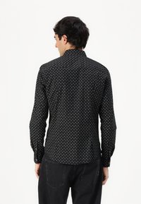 Schwarzes Hemd mit einem geometrischen weißen Muster, langen Ärmeln und einem Kragen. Der Stoff wirkt glatt und leicht.