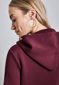 Felpa con cappuccio color borgogna con una texture morbida, che mostra parte della schiena, caratterizzata da un dettaglio di cucitura e un grande orecchino ad anello d'argento.