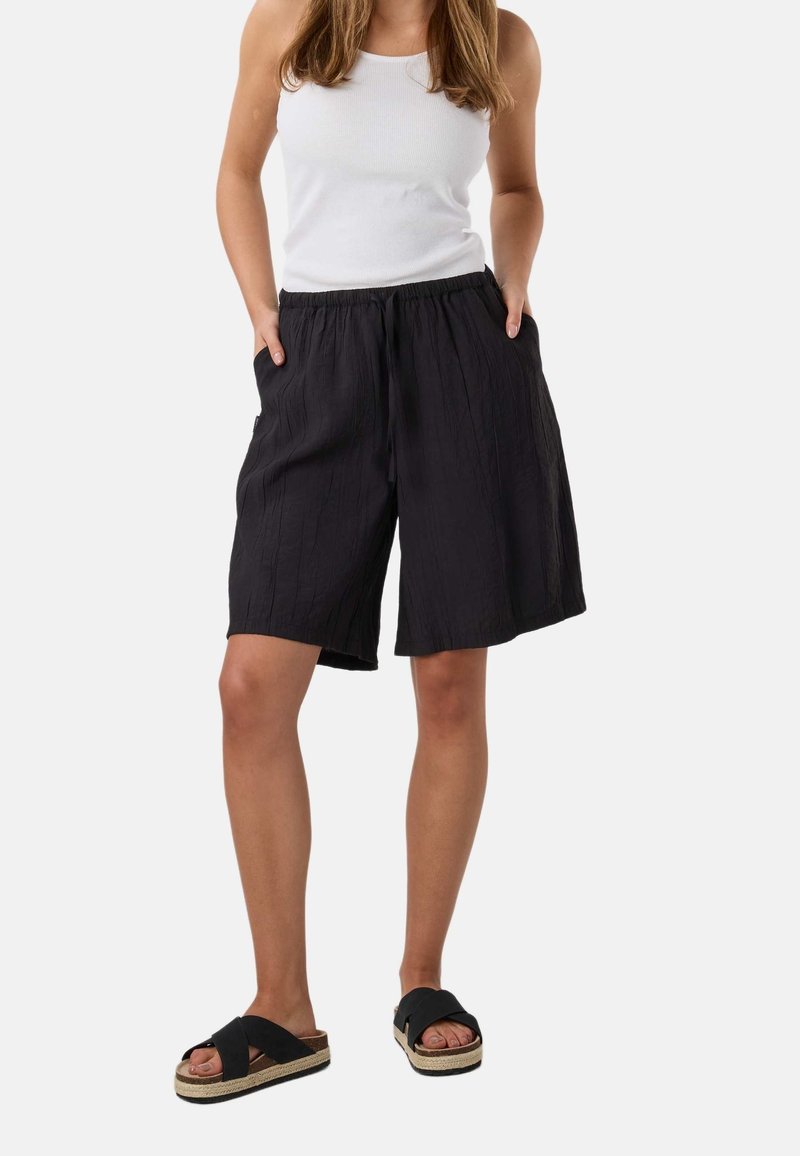 Femme portant un haut blanc sans manches, un short noir ample jusqu'aux genoux avec des poches, et des sandales noires à enfiler avec des semelles épaisses.