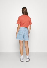 Luźna koszulka koloru rdzawego, zestawiona z jasnoniebieskimi, postrzępionymi szortami jeansowymi. Stylizację dopełniają białe skarpetki i wielokolorowe sneakersy.