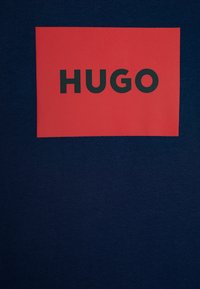 Námořnická modrá tričko s obdélníkovou červenou nášivkou a textem "HUGO" tučným černým písmem, vyrobené z bavlněné tkaniny s hladkou strukturou.