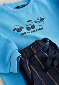 Lichtblauwe sweatshirt met geribde manchetten en een zwart boerderijthema print. Donkerblauwe sweatpants met verticale oranje strepen en een elastische tailleband.