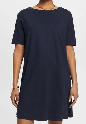 Vrouw in een marineblauwe, korte mouw, loszittende casual jurk met gouden armband en ketting tegen een effen achtergrond.