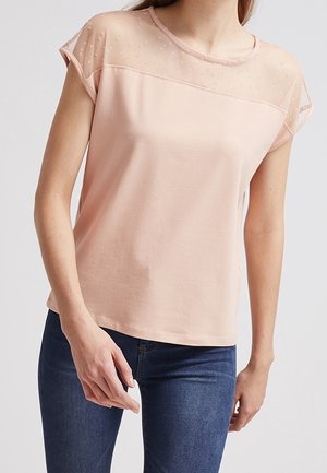 Lyserød kortærmet top med en gennemsigtig prik-mønstret overdel ved halsen, blødt stof og afslappet pasform. Båret med mørkeblå jeans.