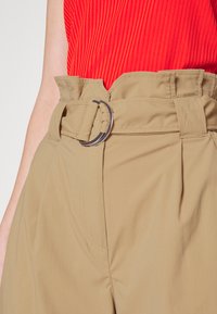 Beige, hoch taillierte Hose mit einem breiten Gürtel und einer silberfarbenen runden Schnalle. Glatter Stoff mit subtilen Nähten.
