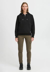 Felpa nera con cappuccio, tasca frontale, zip e accessori in argento, abbinata a pantaloni cargo color oliva e sneakers nere.