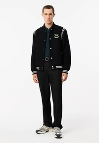 Schwarze Collegejacke mit weißen Streifen, grünem Lacoste-Logo, getragen über einem dunkelgrünen Poloshirt, kombiniert mit schwarzen Hosen und grauen Sneakers.