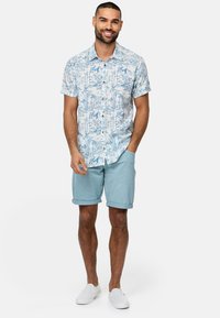 Camisa de manga corta azul y blanca con un diseño de palmeras, combinada con pantalones cortos azul claro y zapatillas blancas sin cordones.