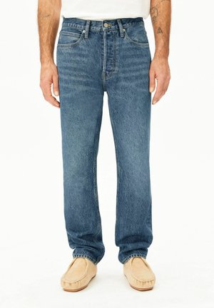 Mann trägt blaue Jeans mit geradem Bein und beige Slip-On Schuhe, steht vor weißem Hintergrund.