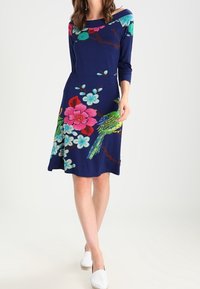 Marinblå klänning med långa ärmar och off-shoulder-design, prydd med livfulla blommiga och fågelmönster i rosa, grönt och turkost.