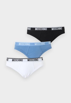 Meztelen lábú férfi modell áll, kék tervezői alsónadrágot visel szürke Moschino derékpánttal, sima fehér háttér előtt.