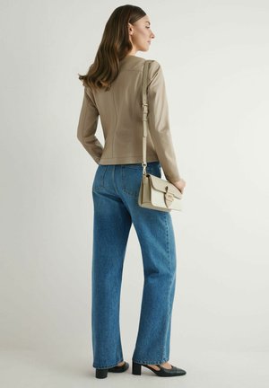 Kobieta w beżowej skórzanej kurtce i niebieskich dżinsach, trzymająca pasującą beżową torebkę, stojąca bokiem na jednolitym tle.