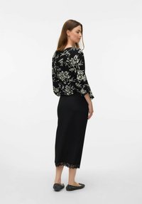 Blouse noire à fleurs avec des manches longues, associée à une jupe crayon noire avec une bordure en dentelle. Le modèle porte des chaussures plates à motif.