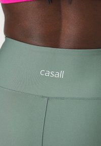 Zelene tajice od glatke tkanine, s ušivenim pojasom i bijelim "casall" logom sprijeda.