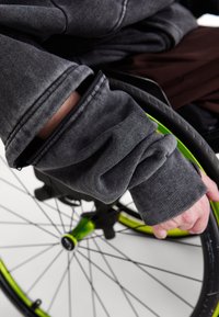 Pelēks satverams džemperis ar ribotu piedurknēm. Redzama roka, kas tur ratiņkrēsla ratu, ar zaļiem akcentiem un plāniem spieķiem.