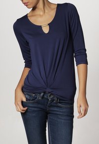 Mörkblå långärmad topp med rund halsringning och en nyckelhålsdetalj, som har en knut vid midjan och en slät tygstruktur.