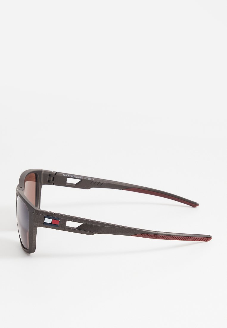 Sonnenbrille mit einem dunkelgrauen Kunststoffrahmen, braun getönten Gläsern und roten Akzenten. Verfügt über geometrische Ausschnitte an den Bügeln. Leichtes Design.