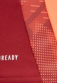 Kurzärmliges Sportoberteil in Rot und Orange, mit strukturierten Mustern und glattem Stoff. Inklusive Logo des Herstellers in Weiß gedruckt.