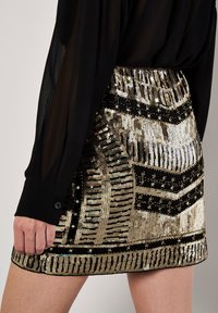 Kurze schwarze Paillettenrock mit goldenen Streifen und geometrischen Mustern. Die Textur ist glänzend und reflektierend, mit einem enganliegenden Bund.