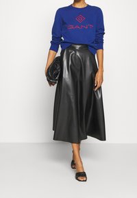 Modrá mikina s červeným logem spárovaná s černou koženou midi sukní a černými plochými sandály. Modelka drží černou kabelku.