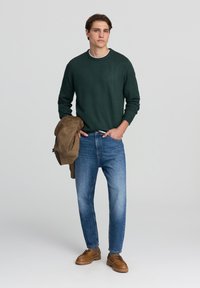 Maglione verde a maglia, jeans blu e scarpe marroni. Il modello tiene un giubbotto beige. Design semplice, vestibilità casual e colori a contrasto.