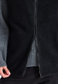 En persons hand håller i kanten på en svart fleecejacka med dragkedja, som bärs över en grå ribbad tröja och svarta byxor.