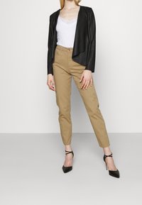 Czarna kurtka z sztucznej skóry z drapowanym przodem, biała bluzka z koronkowym wykończeniem, beżowe slim-fit spodnie oraz czarne szpilki. Neutralne tło.