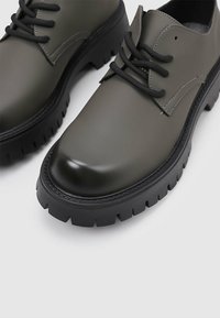 Zapatos de cuero verdes con una suela negra gruesa, puntera redonda y cordones negros; acabado suave con detalles de costura tonales.